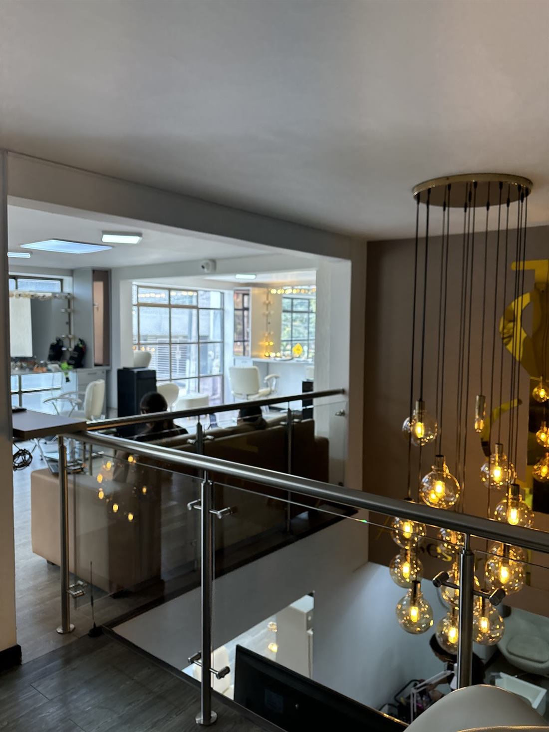 Mezzanine del salón con baranda de vidrio, sillas blancas y lámpara de bombillos colgantes